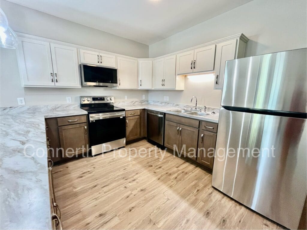 Modern kitchen with stainless steel appliances, including a fridge and oven. Light and dark wood cabinets, marble countertops, and wood flooring. Text on image: Connerth Property Management.