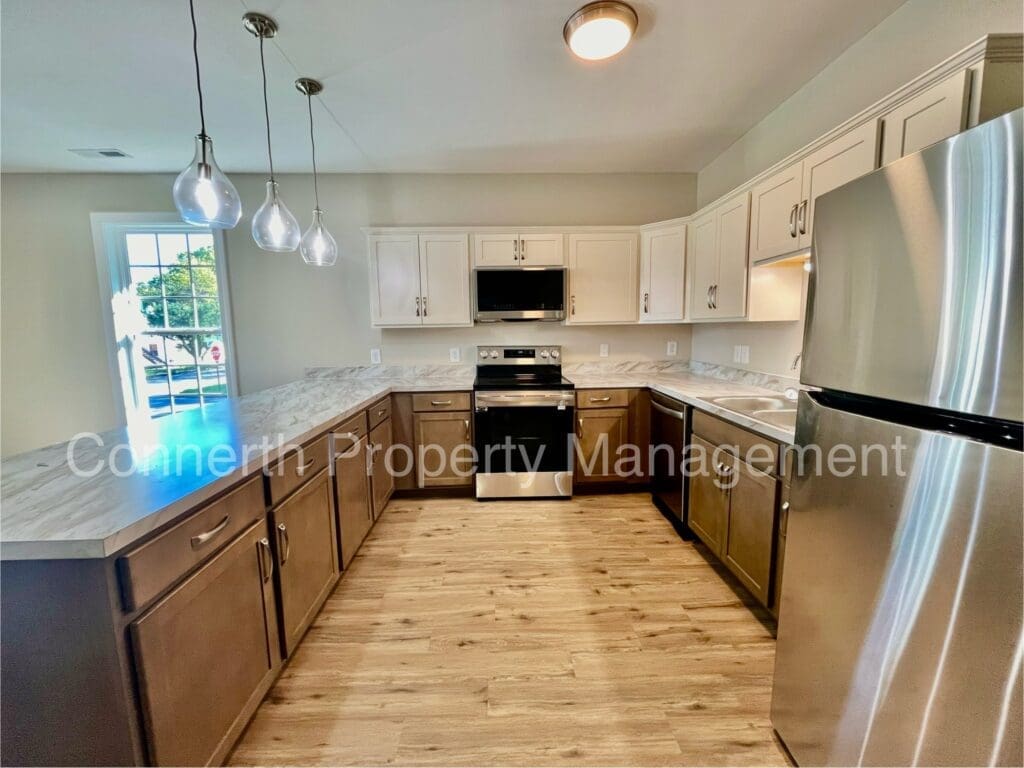 A modern kitchen with stainless steel appliances, wood flooring, light-colored countertops, dark wood lower cabinets, and white upper cabinets. Hanging pendant lights illuminate the area.