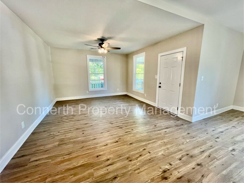 Empty room with wood flooring, beige walls, ceiling fan with lights, two windows, and a white door. Text watermark present.