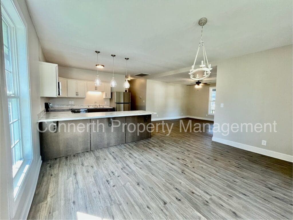 Spacious kitchen and dining area with wooden floors, modern lighting, a large island, and white cabinets. Stainless steel appliances are visible in the background.