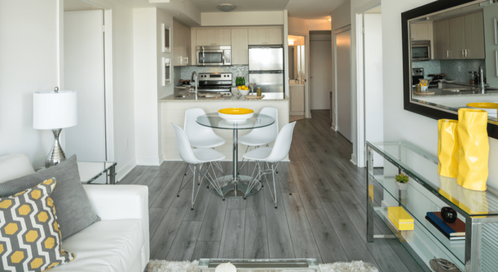 Modern, bright apartment interior with a glass dining table, white chairs, gray flooring, and neutral decor, including yellow accents. Kitchen with stainless steel appliances is visible in the background. Renting in Woodridge Estates, Clarksville TN