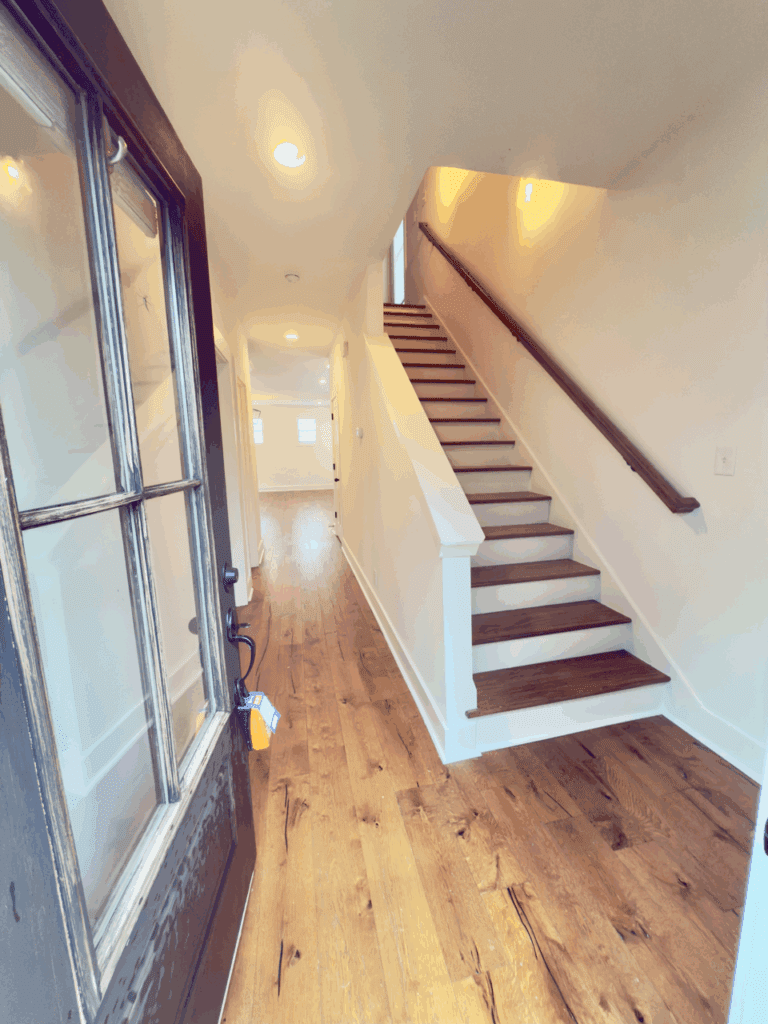 A view from an open front door showing a hallway with wooden floors, white walls, and a staircase leading to the upper floor.