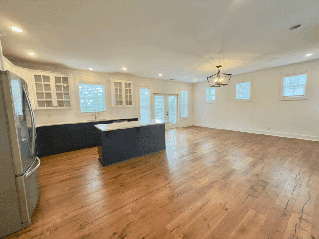 Spacious empty kitchen and living area with wood floors, white cabinets, center island, stainless steel appliances, and multiple windows.