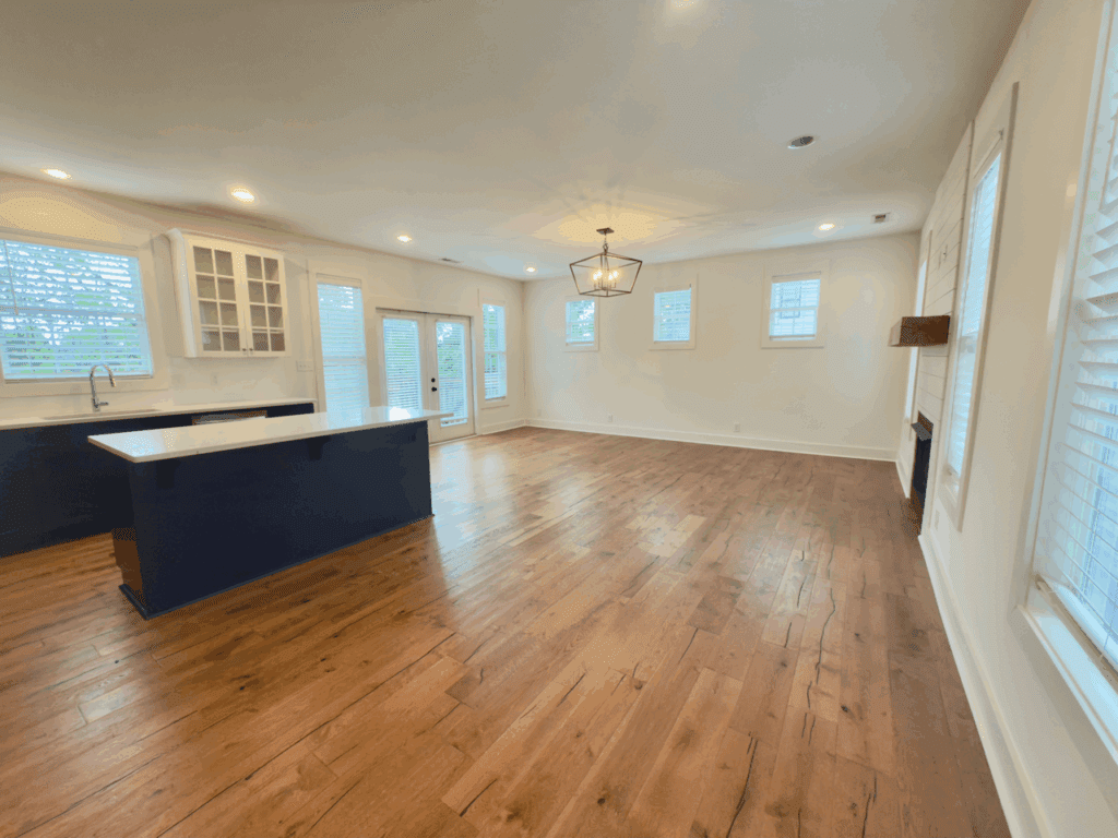 Open-concept kitchen and living area with hardwood floors, white walls, large windows, a kitchen island, and a light fixture above the living space.