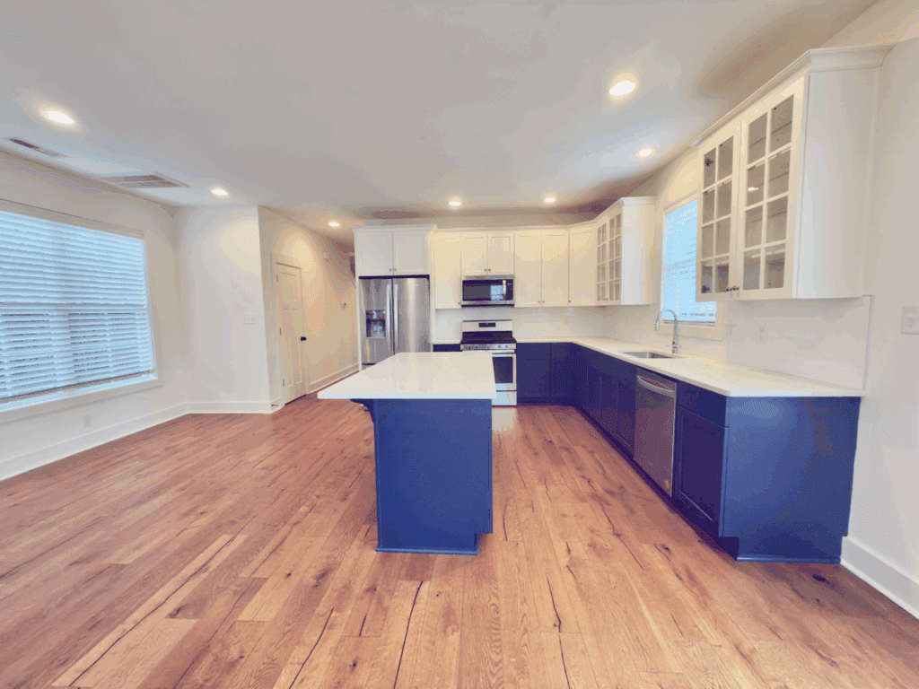 Modern kitchen with white upper cabinets, navy blue lower cabinets and island, stainless steel appliances, hardwood floor, and large windows.