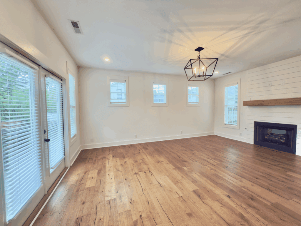 Empty living room with hardwood floors, white walls, a modern chandelier, fireplace, and multiple windows; glass doors lead outside.