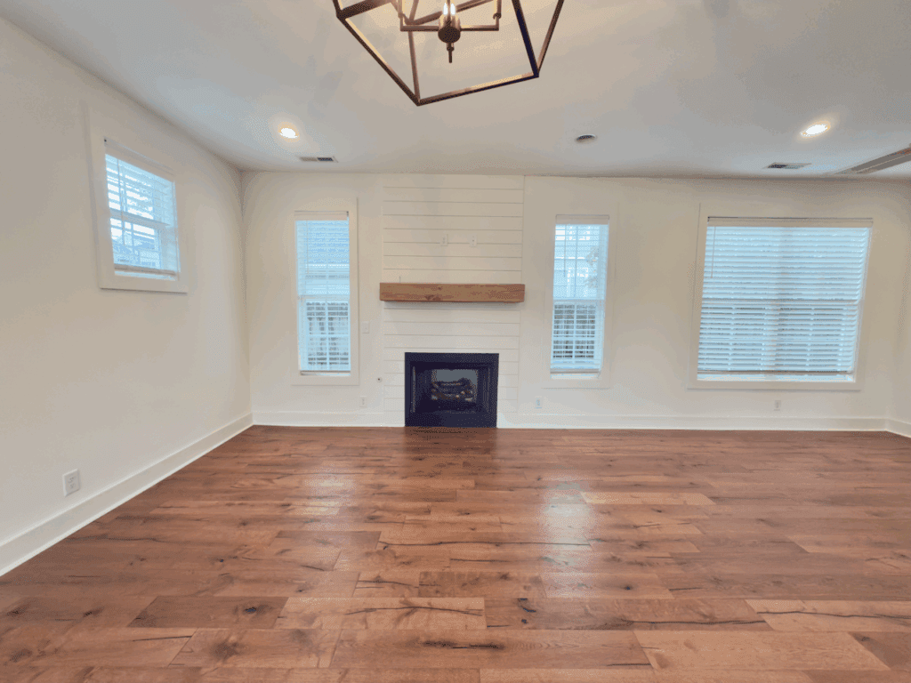 An empty living room with hardwood floors, three windows, white walls, a black fireplace with a wooden mantel, and a modern ceiling light fixture.