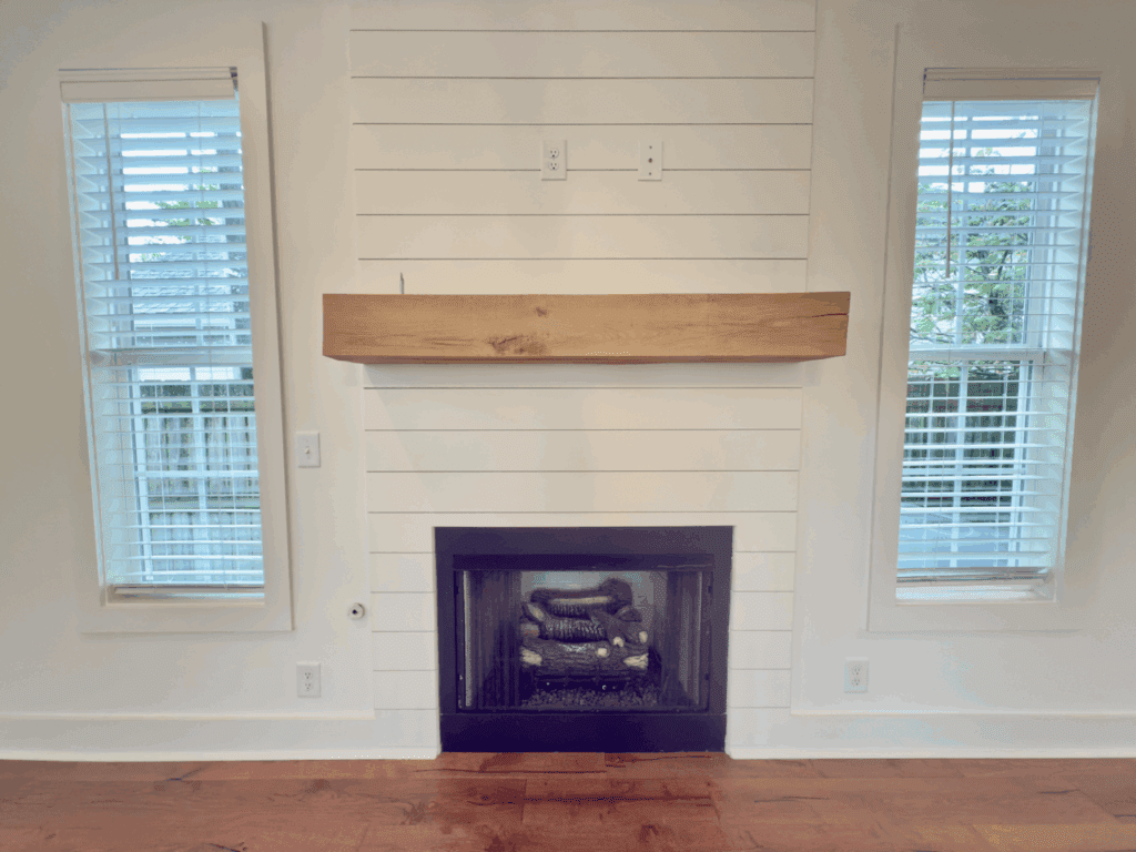 A modern fireplace with a wooden mantel is set into a white shiplap wall between two windows with white blinds; electrical outlets are above the mantel.