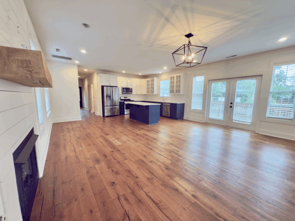 Open-concept living area with hardwood floors, a modern kitchen with white cabinets and stainless appliances, large windows, and double glass doors leading outside.