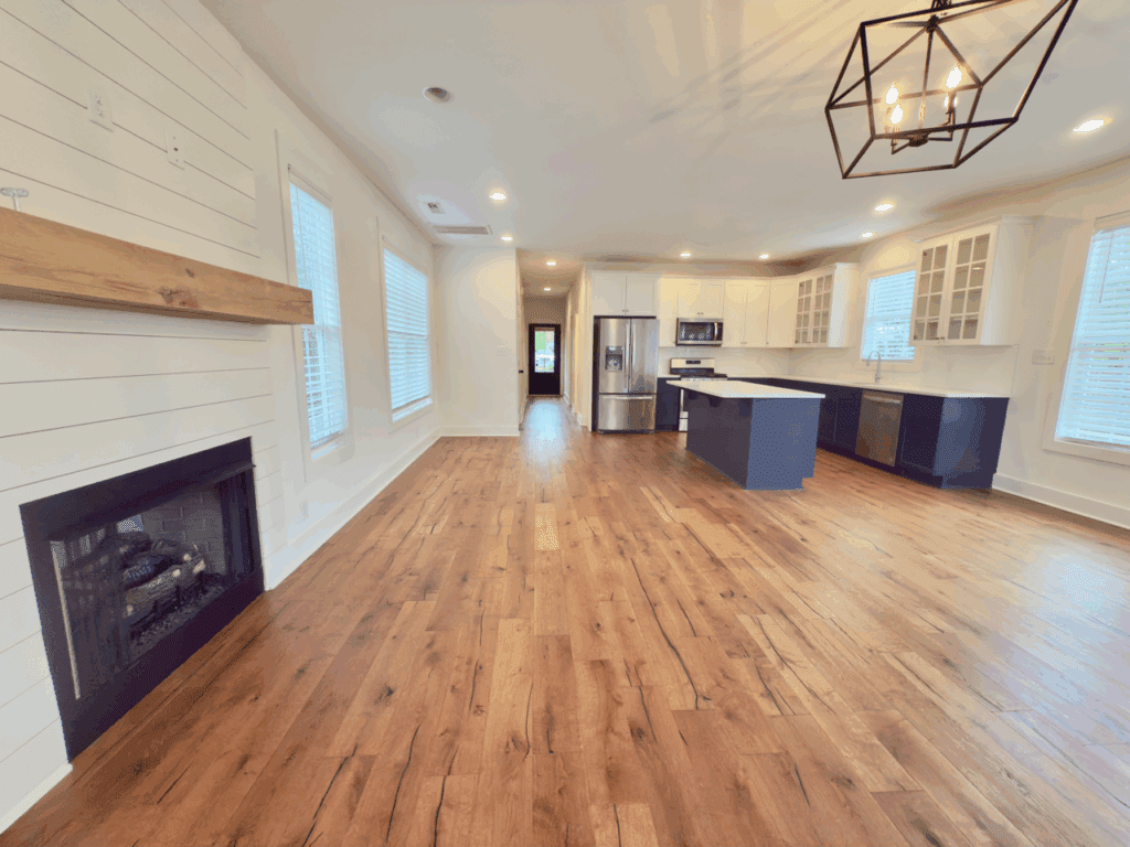 Open-concept kitchen and living area with hardwood floors, white cabinetry, blue island, modern light fixture, fireplace with wood mantel, and large windows.