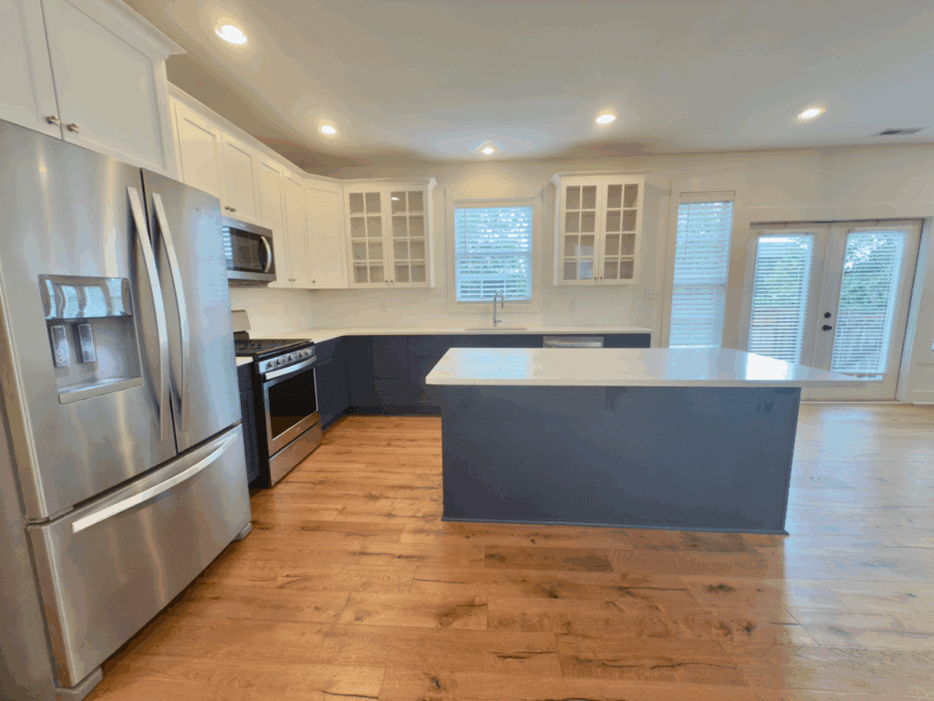 Modern kitchen with stainless steel appliances, white upper cabinets, glass cabinet doors, blue island, and hardwood floors; large windows and a glass door let in natural light.