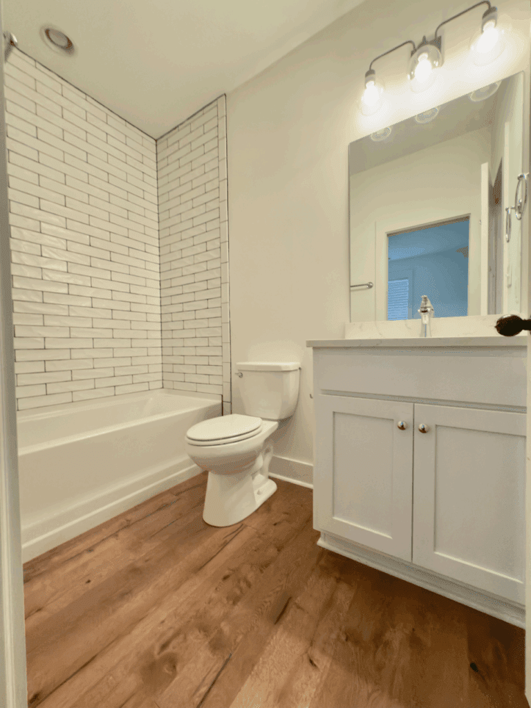 Modern bathroom with white subway tile shower, white toilet, white vanity with sink and mirror, and wood flooring under bright light fixtures.