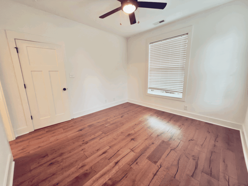 Empty room with white walls, a wooden floor, a window with blinds, a ceiling fan with light, and a closed white door.