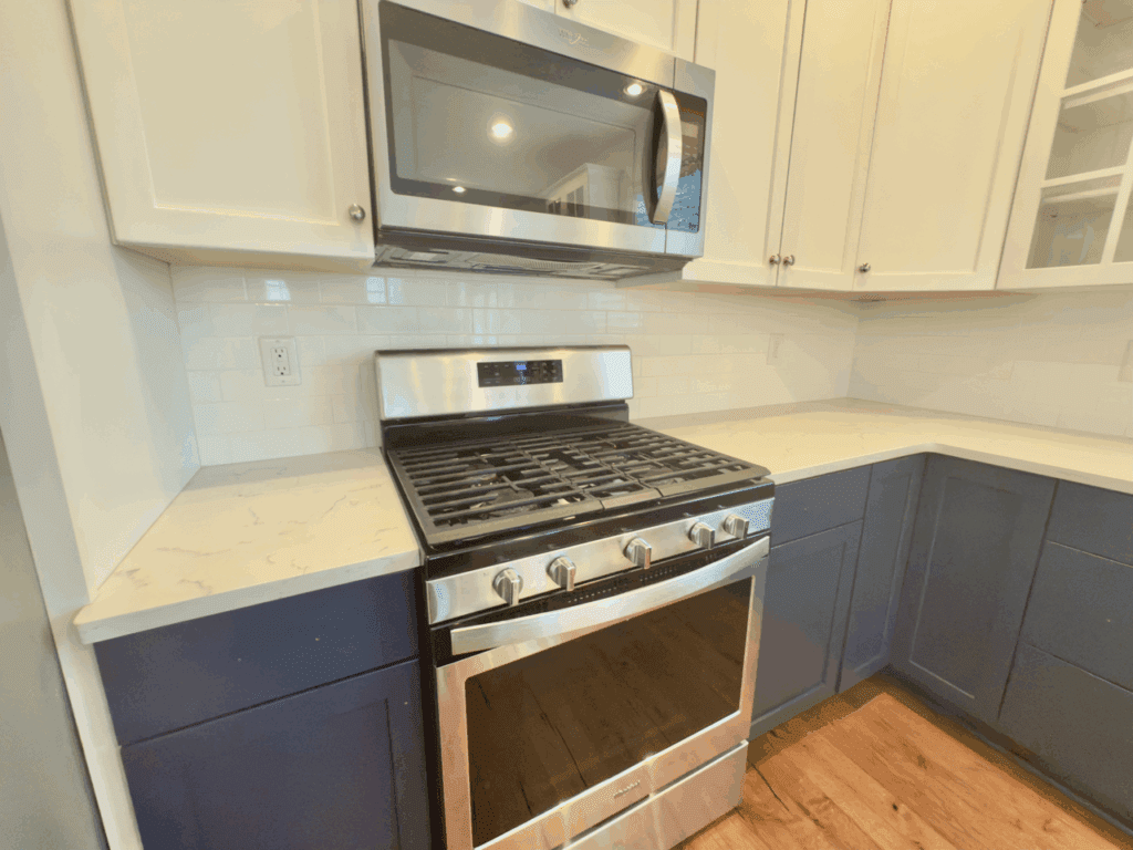 Modern kitchen with a stainless steel gas stove and microwave, white upper cabinets, dark blue lower cabinets, and white quartz countertops.