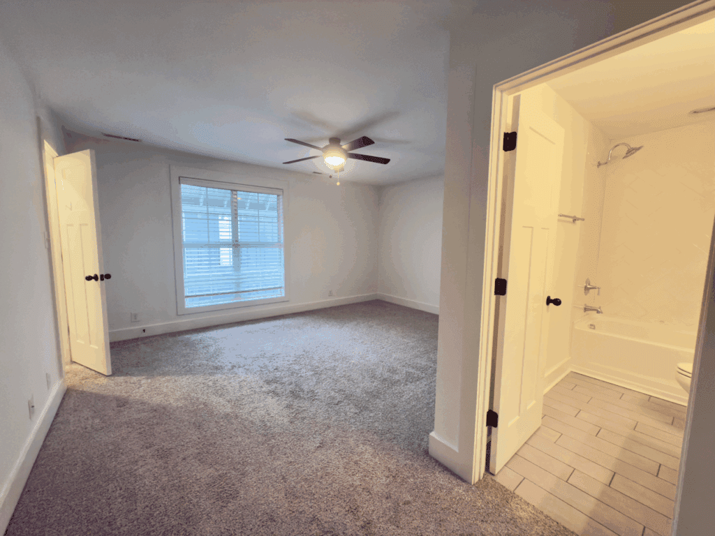 Carpeted bedroom with a ceiling fan, large window with blinds, and adjoining bathroom with tiled floor and bathtub.