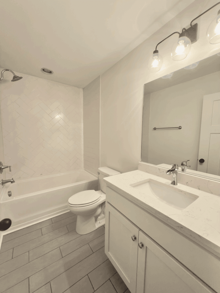 Modern bathroom with white vanity, marble countertop, toilet, bathtub with shower, herringbone tile wall, and large mirror under three-bulb light fixture.