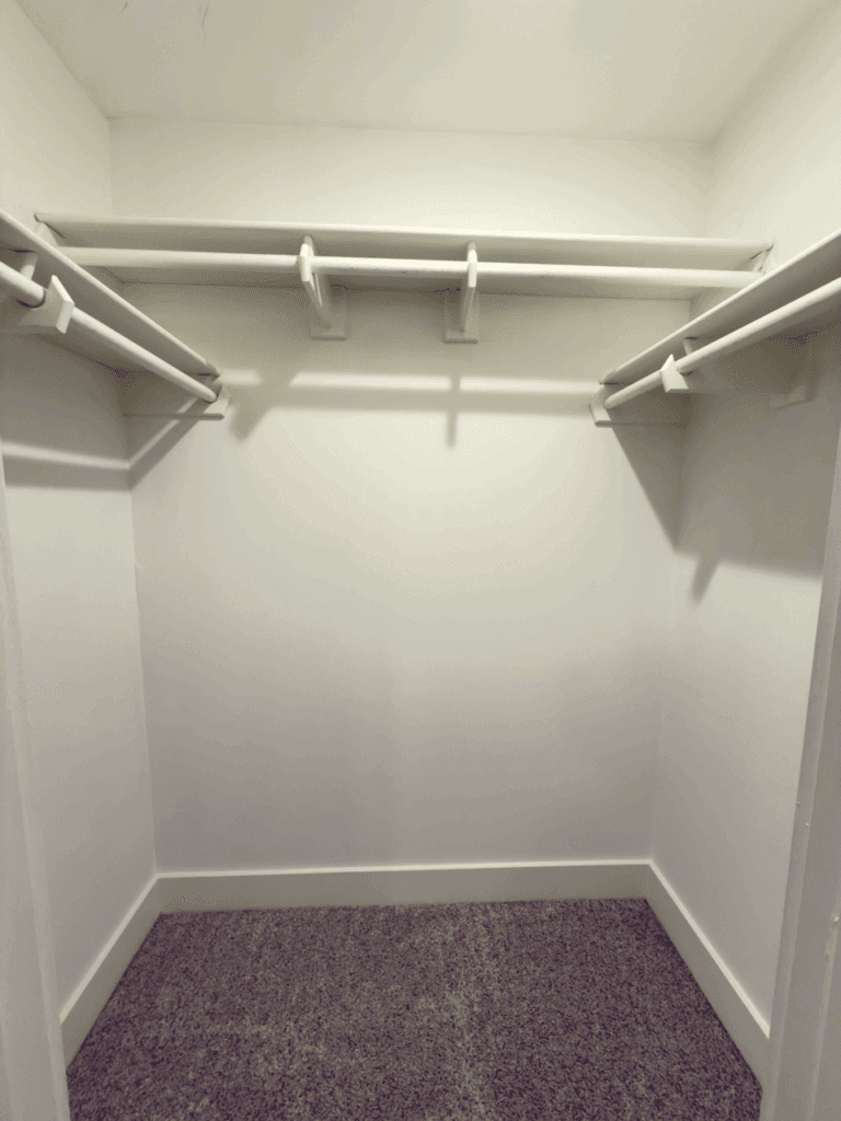 Empty walk-in closet with white walls, three wooden hanging rods, and a speckled carpeted floor.