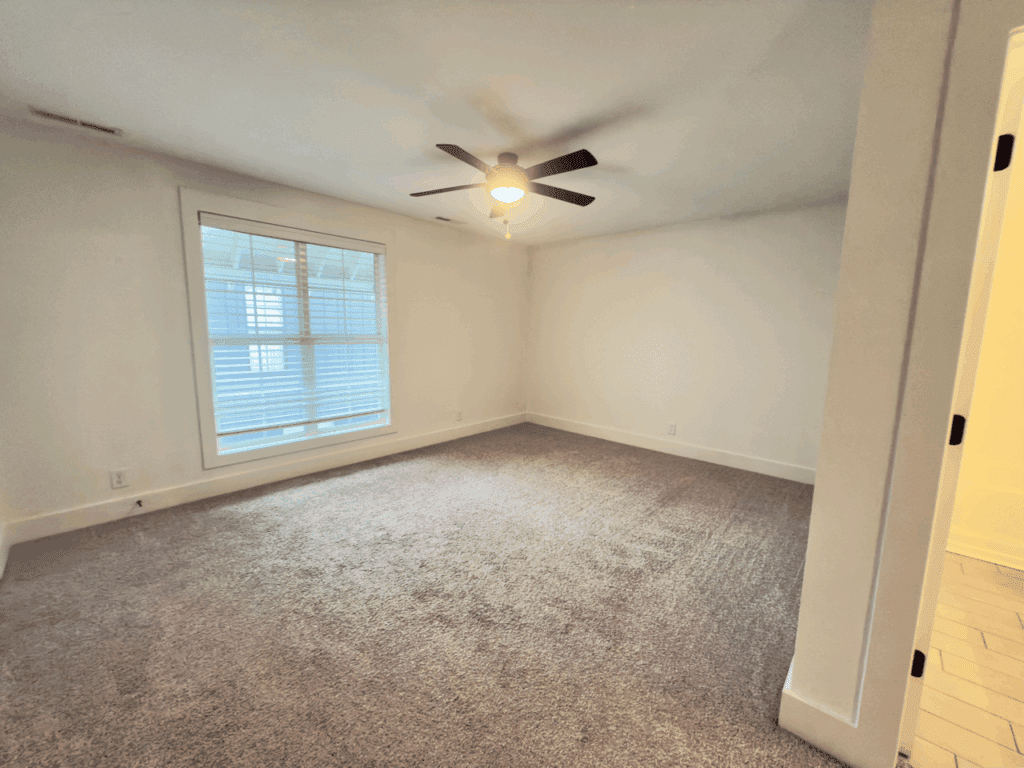 Empty room with beige carpet, white walls, a large window with blinds, a ceiling fan with light, and a partially visible doorway leading to a bathroom.