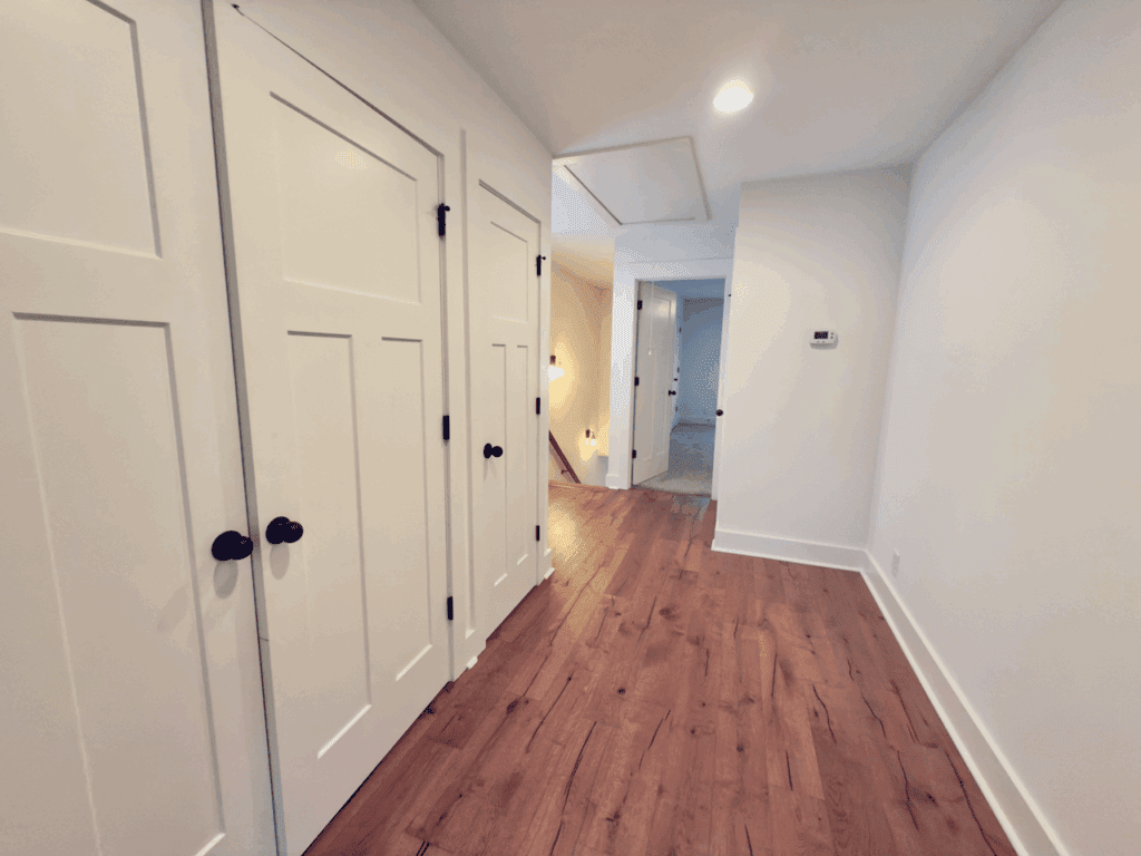 A hallway with wooden floors, white walls, and three closed white doors on the left; recessed ceiling lights are visible.