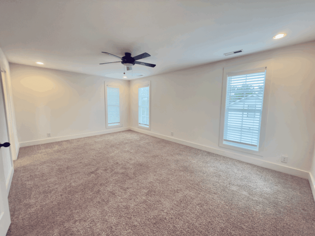 Empty, carpeted room with white walls, two tall windows, a ceiling fan with lights, and recessed lighting.