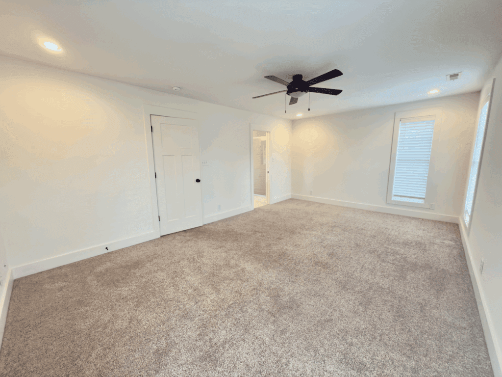 A spacious, empty room with light-colored walls, beige carpet, recessed ceiling lights, a ceiling fan, two windows, and a white door leading to an adjacent room.