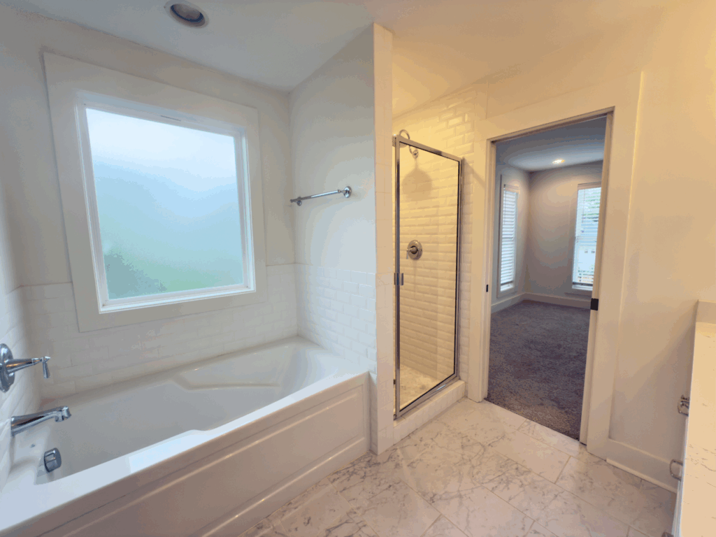 A bathroom with a bathtub, a glass shower enclosure, white tile walls, marble floor, and a window, connected to an adjacent carpeted room.