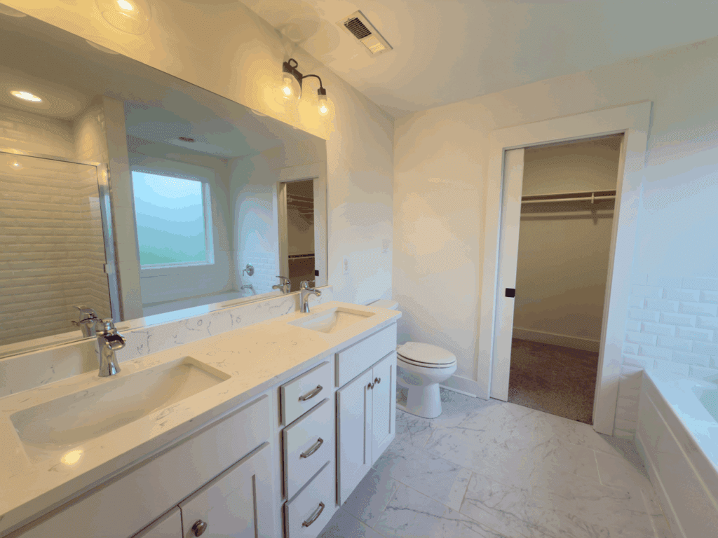 Modern bathroom with double sinks, large mirror, white cabinetry, toilet, walk-in closet, and a glimpse of a bathtub and tiled shower.