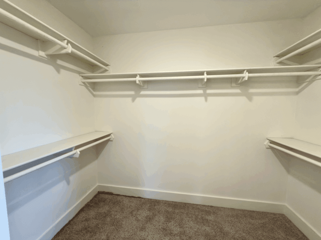 Empty walk-in closet with white shelves and rods on three walls, and beige carpeted floor.