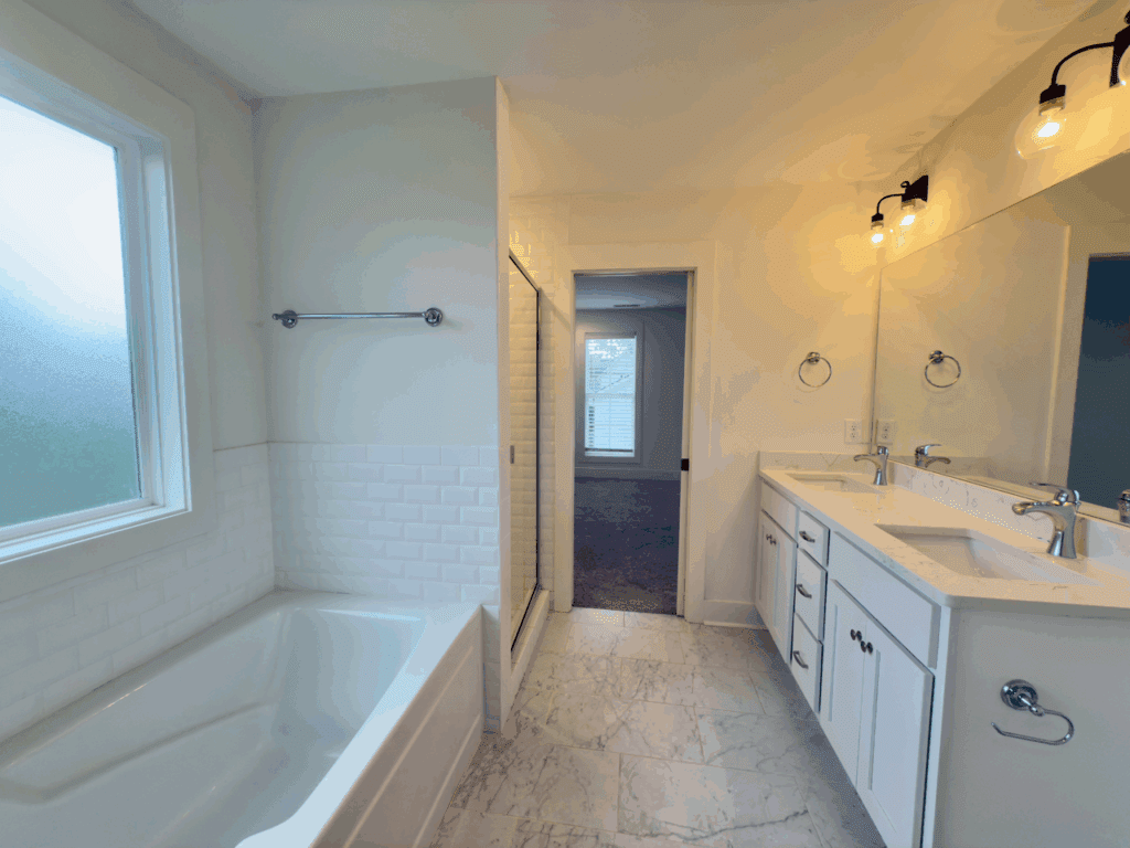 Modern bathroom with a bathtub, walk-in shower, double sink vanity, marble floor, and frosted window. Bright lighting and white cabinetry are visible.