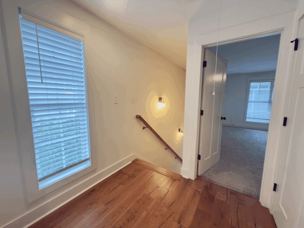 A hallway with hardwood floors leads to a carpeted room, featuring windows with blinds and a staircase with a handrail and wall-mounted lights.