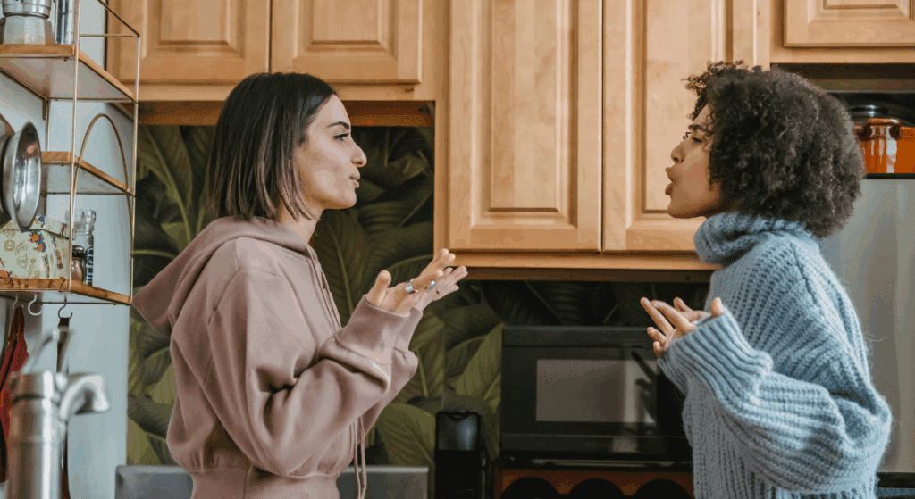 Two women stand in a kitchen facing each other, engaged in a heated conversation with expressive hand gestures.