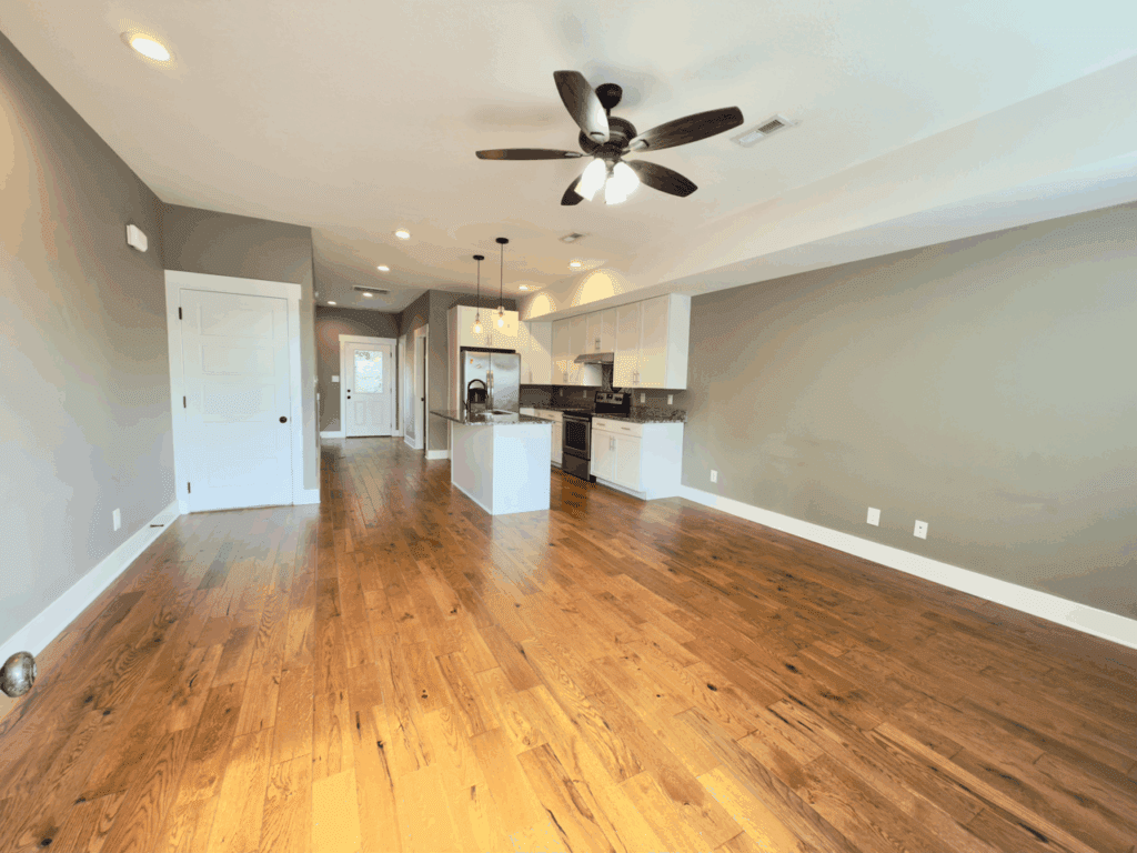 Open-plan kitchen and living area with wooden floors, gray walls, white cabinets, ceiling fan, and recessed lighting; natural light enters through a back door.