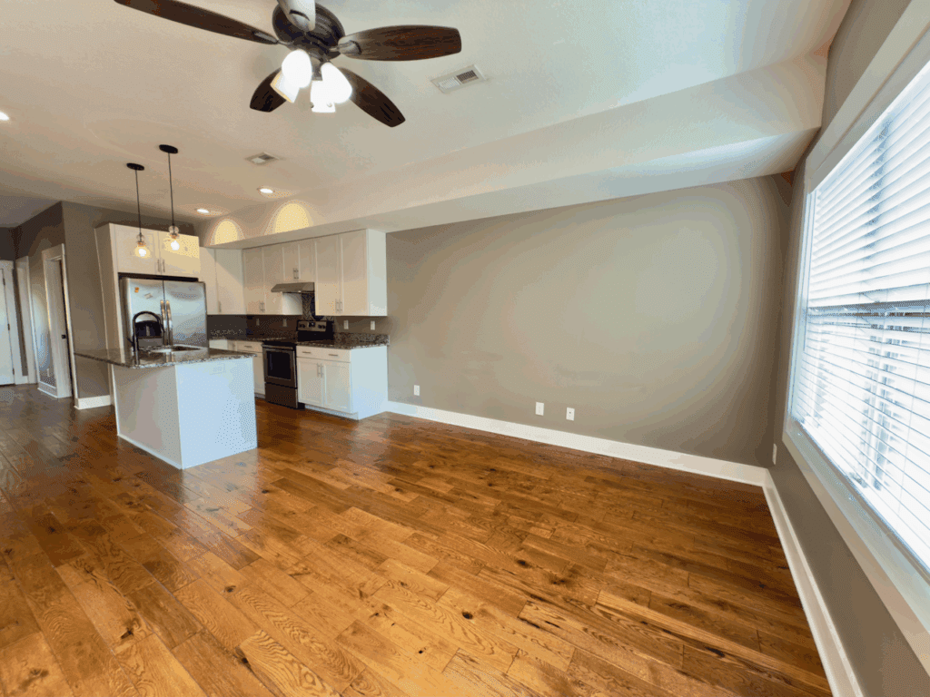 Open-concept kitchen and living area with hardwood floors, stainless steel appliances, white cabinets, neutral walls, and large windows with blinds.