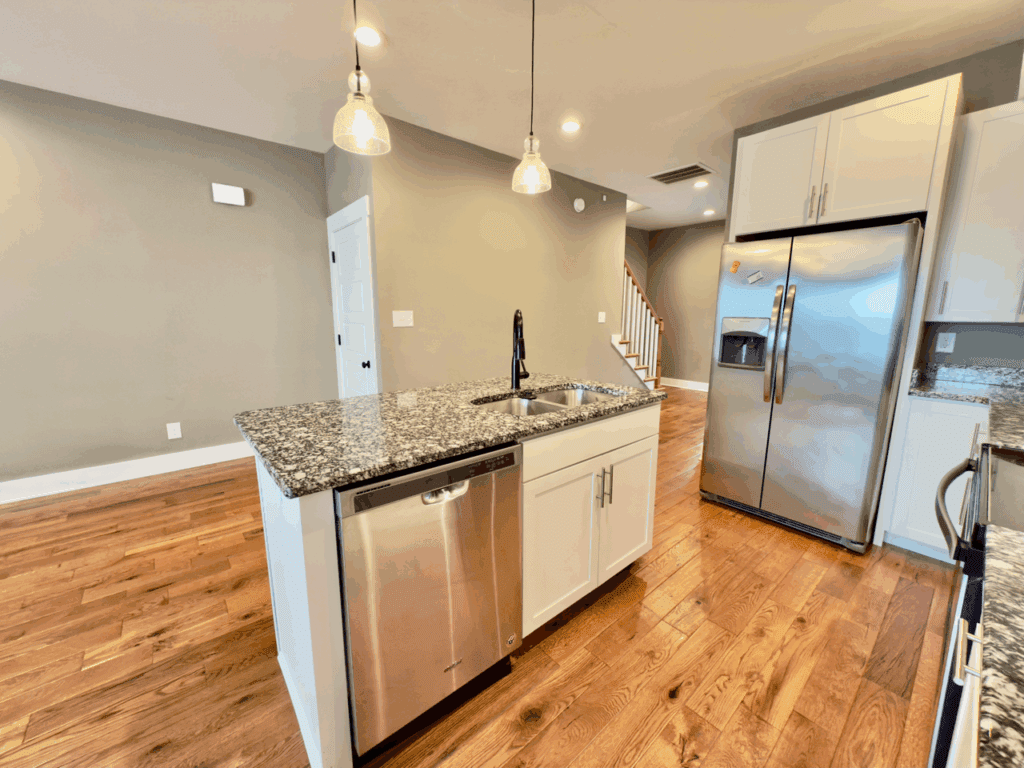 Modern kitchen with stainless steel appliances, granite island with sink and dishwasher, white cabinets, and hardwood floors. Stairs and gray walls are visible in the background.