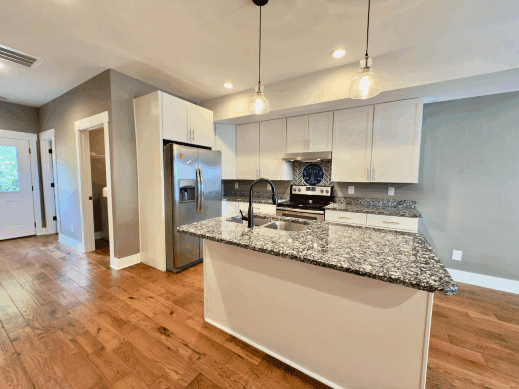Modern kitchen with stainless steel appliances, granite countertops, white cabinets, an island with a black faucet, pendant lights, and hardwood floors.