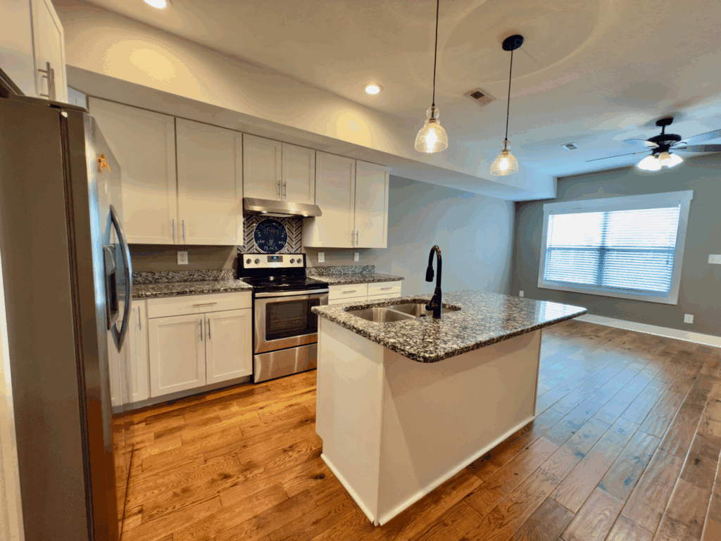 Modern kitchen with stainless steel appliances, white cabinets, granite countertops, center island with sink, pendant lights, and adjacent living area with wood flooring and large window.