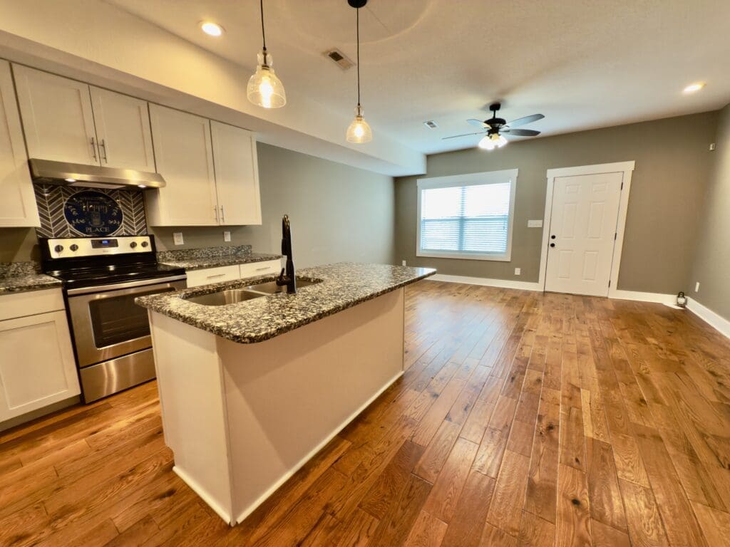 Modern kitchen with granite countertops, stainless steel appliances, wood floors, and an island, open to a living area with a window, ceiling fan, and white entry door.