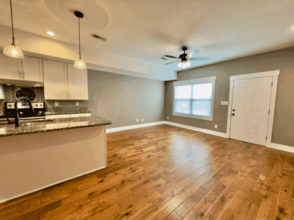 Open-concept room with hardwood floors, kitchen area with granite countertops and pendant lights, and a living space with a ceiling fan and large window.