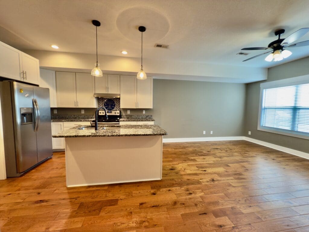 Modern kitchen with granite island, stainless steel appliances, pendant lights, and hardwood floors next to an open living area with ceiling fan and large window.