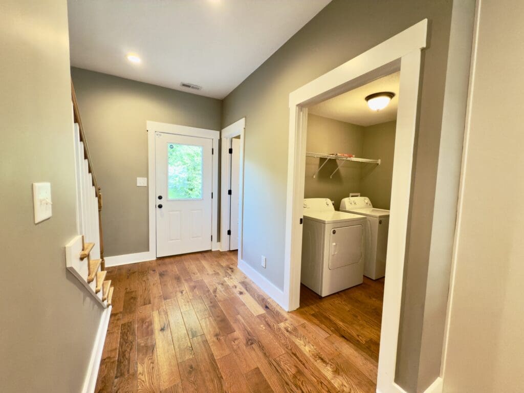 A hallway with wood flooring shows a staircase, a white door with glass panels, and a laundry area with a washer and dryer.