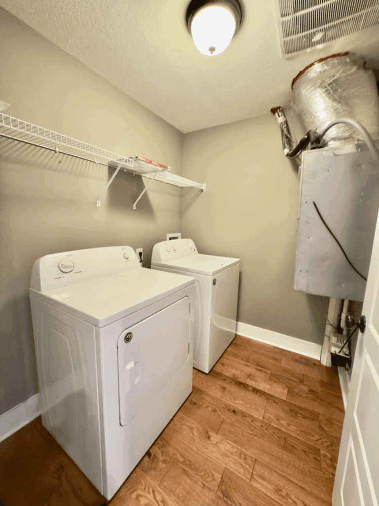 Laundry room with a white dryer and washer, wood floor, wire shelf above, and HVAC unit in the corner against beige walls.