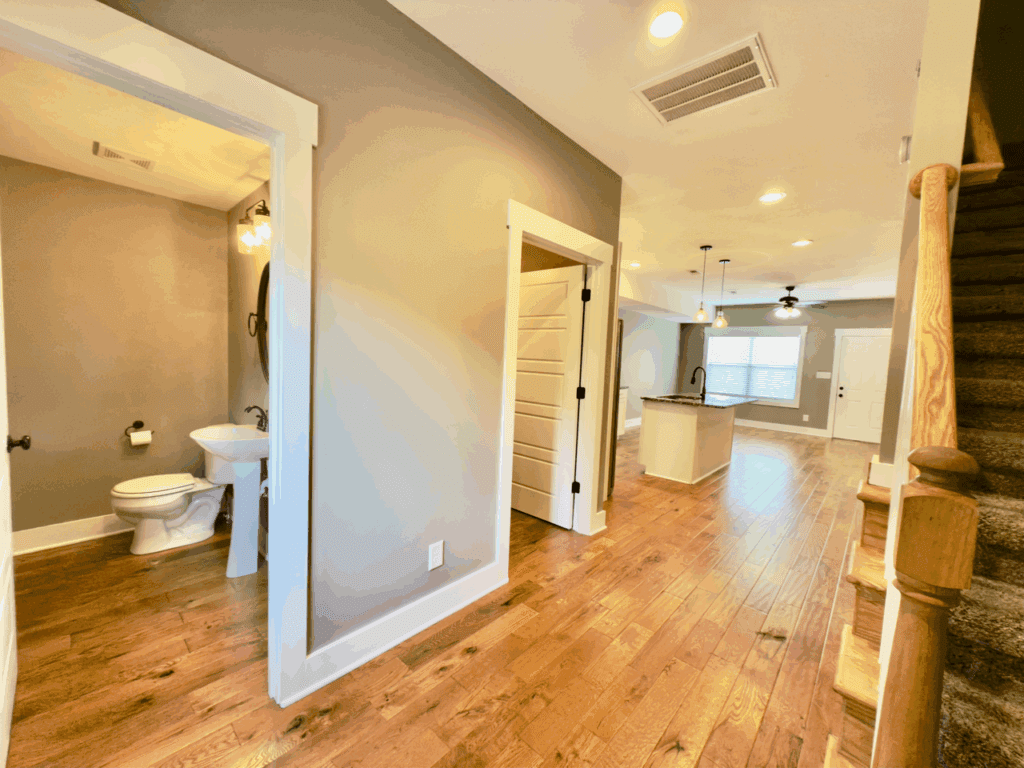 Hallway with wood floors, a staircase to the right, a bathroom on the left, and an open kitchen and living area in the background.