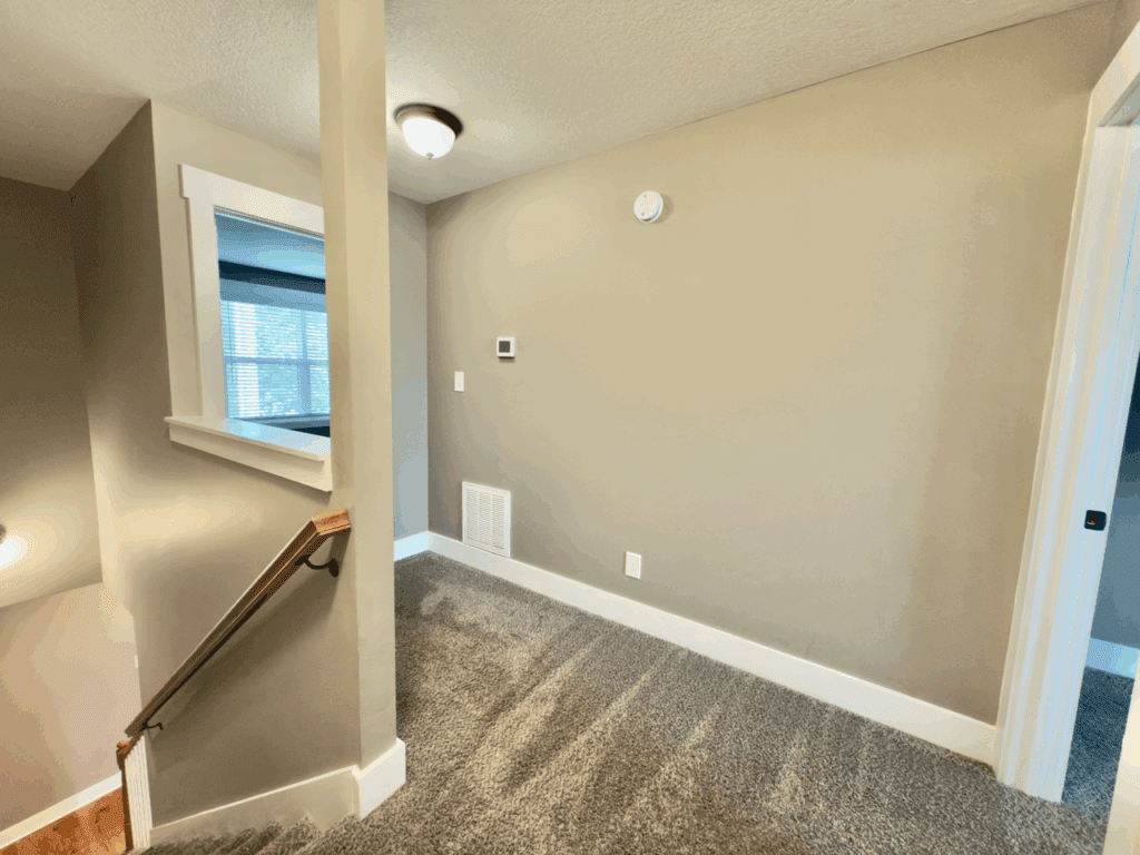 A carpeted hallway with beige walls, white trim, a stair railing, and a small window above the stairs. A thermostat and smoke detector are mounted on the wall.