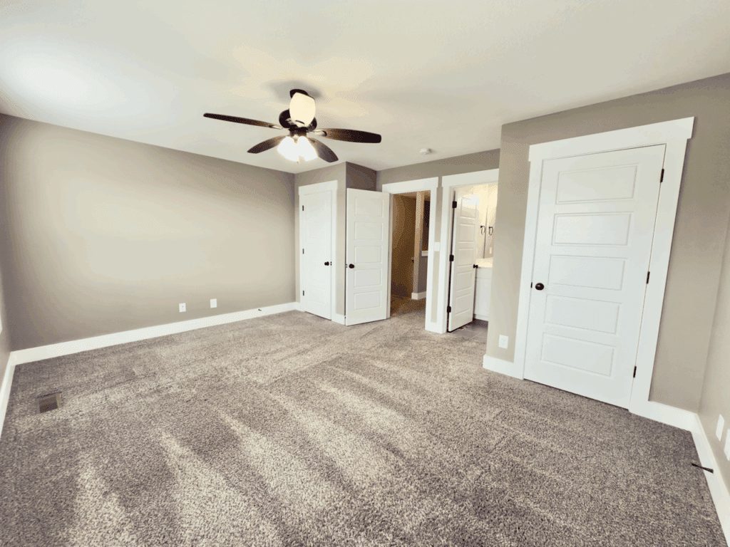Empty carpeted bedroom with beige walls, a ceiling fan with lights, and three white doors, one open to a bathroom. Natural light enters from a window on the left.