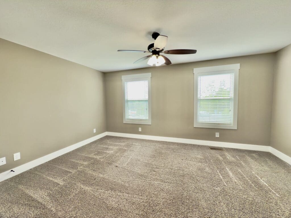 Empty carpeted room with beige walls, two windows with white blinds, white baseboards, and a ceiling fan with light.