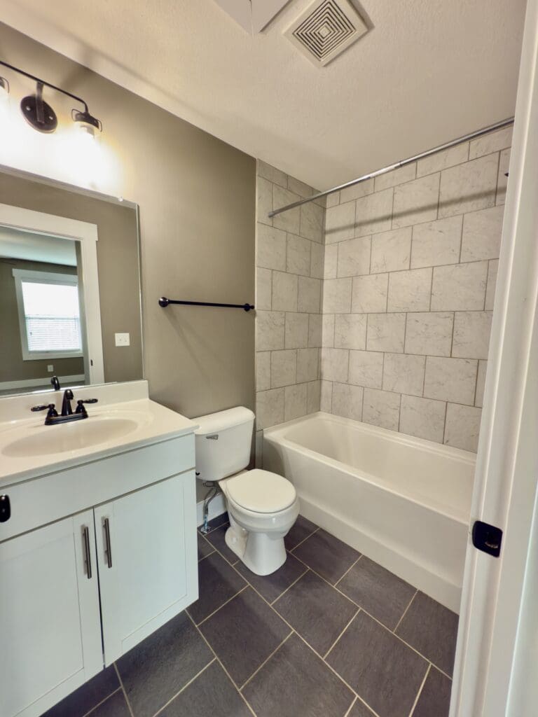 Modern bathroom with white vanity, sink, and toilet next to a bathtub with gray tile walls; large mirror and dark fixtures; gray tile floor.