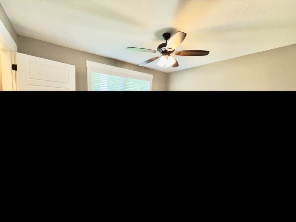Empty bedroom with beige walls, carpeted floor, a window with blinds, ceiling fan with light, and a white door partially open.