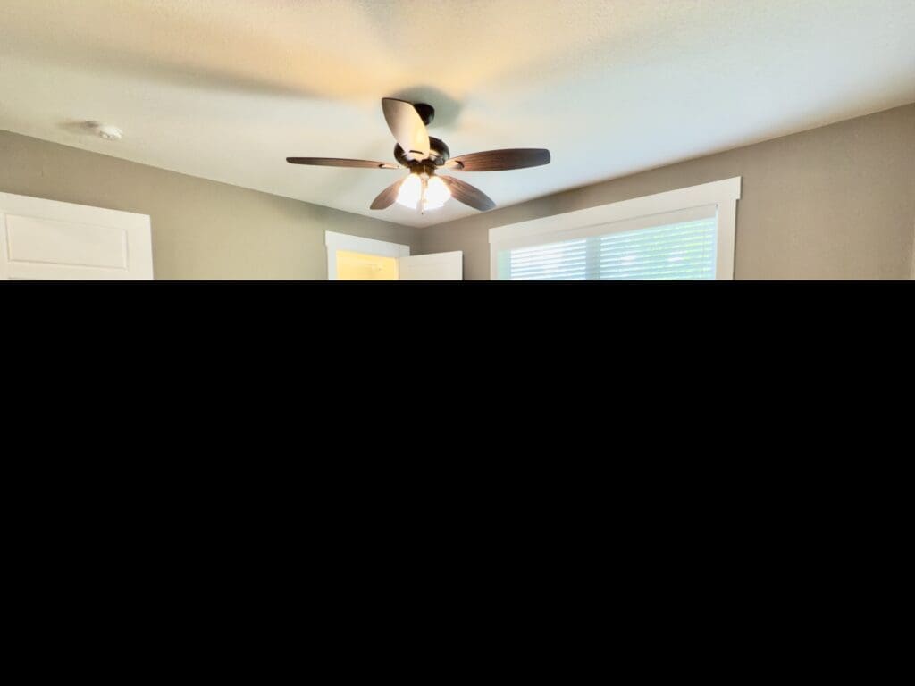 Empty bedroom with gray carpet, beige walls, a ceiling fan with lights, a large window with blinds, and an open door leading to a walk-in closet.
