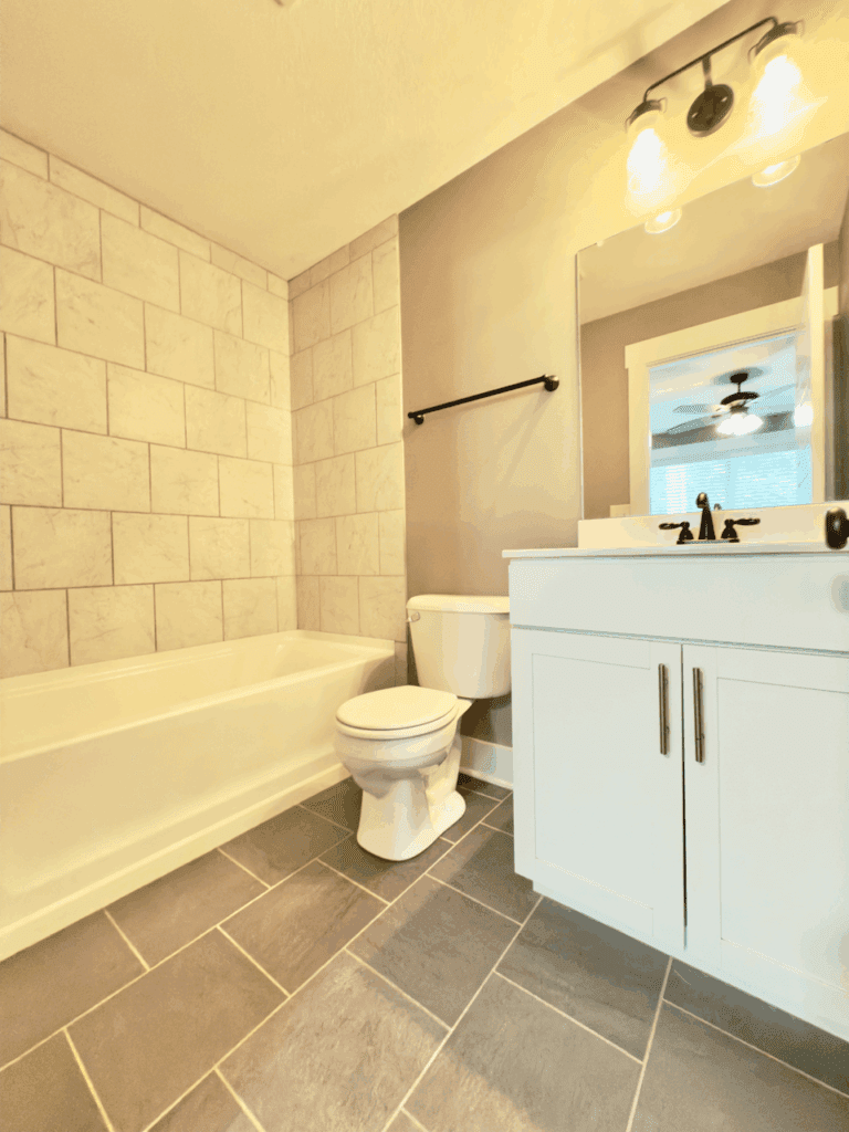 A modern bathroom features a white bathtub, a toilet, a white vanity with sink, a wall mirror, and gray tiled floor and wall.