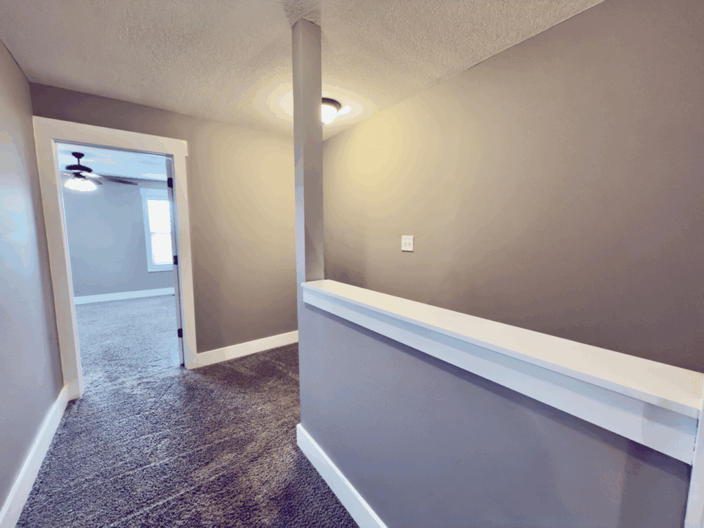 A small carpeted hallway with beige walls, a white trim, a dividing half-wall, ceiling lights, and an open doorway leading into a bedroom with a ceiling fan.